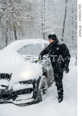 Man dressed in black winter clothing is clearing snow from a car parked in a snowy landscape with trees covered in snow during a winter storm Man dressed in black winter clothing is clearing snow from a car parked in a snowy landscape with trees covered in snow during a winter storm 140031705