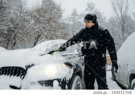 Man in black winter clothing is clearing snow from a car hood with a snow shovel in a snowy outdoor environment surrounded by trees and other vehicles Man in black winter clothing is clearing snow from a car hood with a snow shovel in a snowy outdoor environment surrounded by trees and other vehicles 140031708