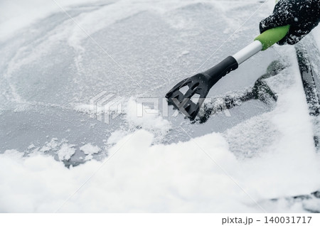 Person using a black and green snow scraper to remove snow from a car windshield covered in white snow during winter weather conditions in a cold environment Person using a black and green snow scraper to remove snow from a car windshield covered in white snow during winter weather conditions in a cold environment 140031717