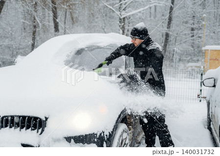 Man in black winter clothing is clearing snow from a car windshield during a snowfall in a wooded area with trees and a fence visible in the background 140031732
