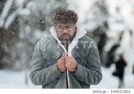 Young man with curly hair wearing a gray winter coat is standing outdoors in the snow, holding his coat closed against the cold while snowflakes fall around him Young man with curly hair wearing a gray winter coat is standing outdoors in the snow, holding his coat closed against the cold while snowflakes fall around him 140031802