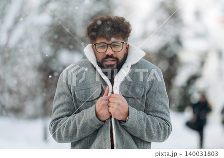 Man with curly hair and glasses is standing outdoors in a snowy landscape, wearing a gray coat with a white collar, appearing cold and bundled up against the winter weather Man with curly hair and glasses is standing outdoors in a snowy landscape, wearing a gray coat with a white collar, appearing cold and bundled up against the winter weather 140031803