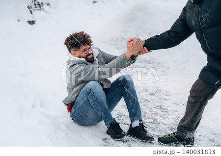 Man with curly hair sitting on snow-covered ground is being helped up by another person in winter clothing on a snowy path during a cold day Man with curly hair sitting on snow-covered ground is being helped up by another person in winter clothing on a snowy path during a cold day 140031813