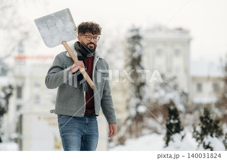 Man with curly hair wearing glasses and a gray jacket is walking outdoors in the snow while holding a shovel, trees and buildings visible in the background during winter Man with curly hair wearing glasses and a gray jacket is walking outdoors in the snow while holding a shovel, trees and buildings visible in the background during winter 140031824