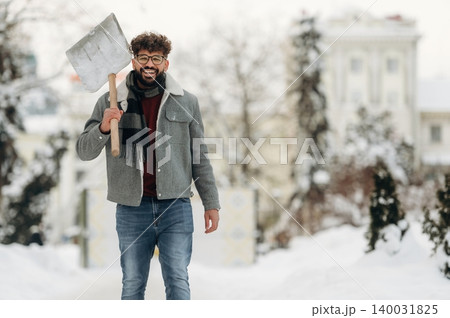 Smiling man with curly hair wearing a gray jacket and scarf is walking through a snowy landscape holding a shovel, trees and buildings visible in the background Smiling man with curly hair wearing a gray jacket and scarf is walking through a snowy landscape holding a shovel, trees and buildings visible in the background 140031825