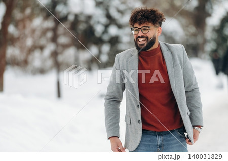 Smiling man with curly hair wearing a gray blazer and red sweater stands in a snowy outdoor setting, surrounded by trees and winter scenery during a cold day Smiling man with curly hair wearing a gray blazer and red sweater stands in a snowy outdoor setting, surrounded by trees and winter scenery during a cold day 140031829