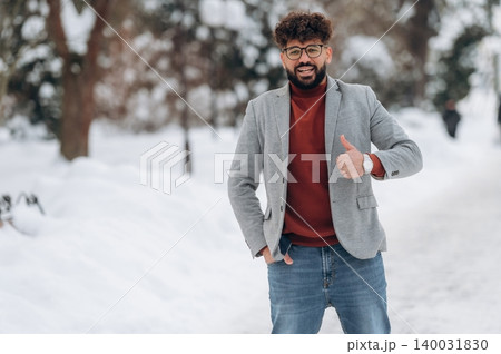Young man with curly hair wearing a gray blazer and red sweater gives a thumbs-up in a snowy park setting with trees and a path visible in the background Young man with curly hair wearing a gray blazer and red sweater gives a thumbs-up in a snowy park setting with trees and a path visible in the background 140031830