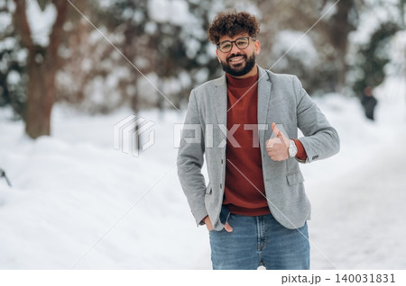 Young man with curly hair wearing a gray blazer and red sweater is standing in a snowy park, smiling and giving a thumbs-up gesture with trees in the background Young man with curly hair wearing a gray blazer and red sweater is standing in a snowy park, smiling and giving a thumbs-up gesture with trees in the background 140031831