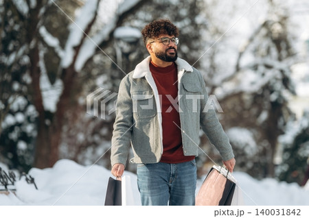 Man with curly hair wearing a gray jacket and red sweater is walking outdoors in winter, holding shopping bags while surrounded by snow-covered trees and a bright sky Man with curly hair wearing a gray jacket and red sweater is walking outdoors in winter, holding shopping bags while surrounded by snow-covered trees and a bright sky 140031842