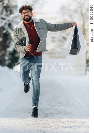Young man with curly hair wearing a gray jacket and red sweater is joyfully running on a snowy path while holding shopping bags in a winter outdoor setting Young man with curly hair wearing a gray jacket and red sweater is joyfully running on a snowy path while holding shopping bags in a winter outdoor setting 140031849