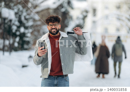 Young man with curly hair wearing a gray jacket and glasses is holding a shopping bag while using a smartphone in a snowy urban setting with blurred pedestrians 140031858