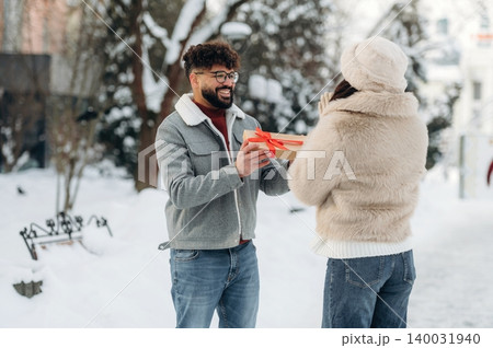 Man with curly hair wearing glasses is giving a gift to a woman in a winter coat during a snowy day in a park with trees and benches in the background Man with curly hair wearing glasses is giving a gift to a woman in a winter coat during a snowy day in a park with trees and benches in the background 140031940