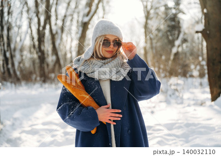 Young woman wearing sunglasses and a winter coat holds a baguette while standing in a snowy forest with trees in the background illuminated by sunlight 140032110