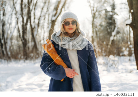 Woman in winter attire holding a baguette stands in a snowy forest, wearing sunglasses and a knitted scarf, with trees and sunlight in the background 140032111