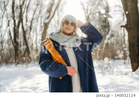 Young woman in winter attire wearing sunglasses and a knitted scarf poses in a snowy forest, sunlight filtering through trees in the background Young woman in winter attire wearing sunglasses and a knitted scarf poses in a snowy forest, sunlight filtering through trees in the background 140032114