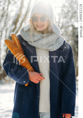 Young woman wearing sunglasses and a warm coat holds fresh baguettes while standing in a snowy outdoor setting with trees in the background Young woman wearing sunglasses and a warm coat holds fresh baguettes while standing in a snowy outdoor setting with trees in the background 140032119