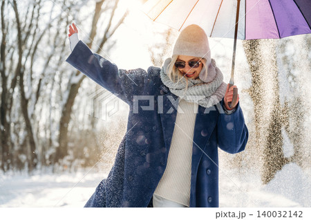 Woman in winter attire joyfully holding a colorful umbrella while standing in a snowy forest, sunlight filtering through trees creating a bright atmosphere 140032142