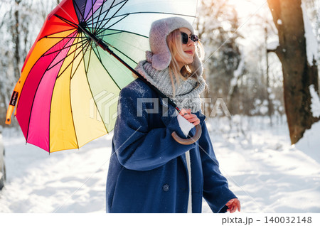 Young woman wearing a blue coat and gray scarf holds a colorful umbrella while walking through a snowy landscape with trees in the background Young woman wearing a blue coat and gray scarf holds a colorful umbrella while walking through a snowy landscape with trees in the background 140032148