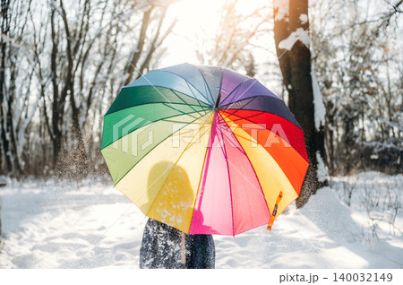 Individual holding a colorful rainbow umbrella in a snowy forest, sunlight filtering through trees, creating a bright and vibrant winter scene Individual holding a colorful rainbow umbrella in a snowy forest, sunlight filtering through trees, creating a bright and vibrant winter scene 140032149