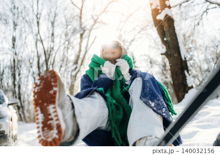 Young woman wearing sunglasses and a green scarf sits in the snow, smiling with sunlight shining through trees in a winter landscape, snow-covered ground and vehicle nearby 140032156