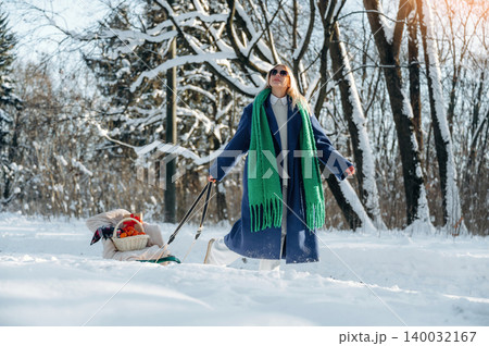 Woman in blue coat and green scarf stands in snowy landscape, pulling sled with colorful blankets and toys, surrounded by trees in winter sunlight Woman in blue coat and green scarf stands in snowy landscape, pulling sled with colorful blankets and toys, surrounded by trees in winter sunlight 140032167