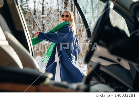 Young woman wearing sunglasses and a blue coat with a green scarf stands outside a car in a snowy forest, smiling and enjoying the winter scenery Young woman wearing sunglasses and a blue coat with a green scarf stands outside a car in a snowy forest, smiling and enjoying the winter scenery 140032181