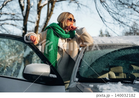 Female model wearing sunglasses and a green scarf leans against a white car with snow on the roof, surrounded by winter trees in a sunny outdoor setting 140032182