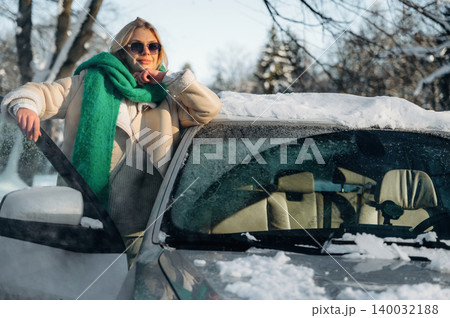 Woman with blonde hair wearing sunglasses and a green scarf stands beside a car covered in snow, winter trees and blue sky visible in the background Woman with blonde hair wearing sunglasses and a green scarf stands beside a car covered in snow, winter trees and blue sky visible in the background 140032188