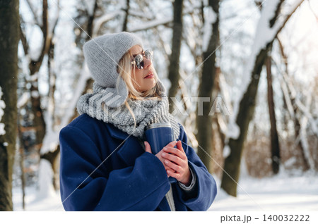 Young woman wearing gray hat and sunglasses holds a thermos while standing in a snowy forest, surrounded by trees covered in snow and sunlight filtering through branches 140032222