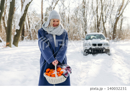 Woman in winter coat and gray hat holds a basket of oranges while standing in snowy landscape with a car parked in the background Woman in winter coat and gray hat holds a basket of oranges while standing in snowy landscape with a car parked in the background 140032231