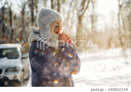 Female figure in gray winter coat and knitted hat stands outdoors in snowy landscape, sunlight creating a soft bokeh effect in the background Female figure in gray winter coat and knitted hat stands outdoors in snowy landscape, sunlight creating a soft bokeh effect in the background 140032239