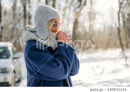 Young woman wearing gray hat and sunglasses warms her hands in winter landscape with snow-covered ground and trees in the background, dressed in blue coat and scarf Young woman wearing gray hat and sunglasses warms her hands in winter landscape with snow-covered ground and trees in the background, dressed in blue coat and scarf 140032241