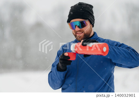 Male hiker wearing blue jacket and sunglasses pours hot drink from red thermos into cup while standing in snowy landscape with blurred trees in background 140032901