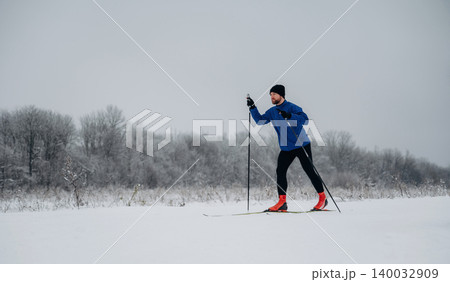 Male cross-country skier in blue jacket and black pants glides through snowy landscape with trees covered in frost under overcast sky Male cross-country skier in blue jacket and black pants glides through snowy landscape with trees covered in frost under overcast sky 140032909