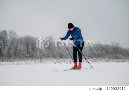 Male cross-country skier in blue jacket and black pants glides over snowy landscape with trees in the background during winter season Male cross-country skier in blue jacket and black pants glides over snowy landscape with trees in the background during winter season 140032912