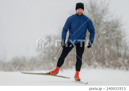 Male cross-country skier in blue jacket and black pants gliding on snow-covered trail with trees in the background during winter season 140032913