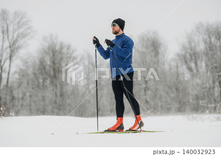 Male cross-country skier in blue jacket and black leggings stands on snow-covered trail, holding ski poles with trees in the background during winter season 140032923