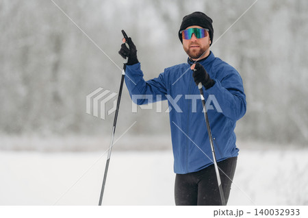 Male cross-country skier wearing sunglasses and blue jacket holds ski poles while standing on snow-covered trail in winter landscape with trees in the background Male cross-country skier wearing sunglasses and blue jacket holds ski poles while standing on snow-covered trail in winter landscape with trees in the background 140032933