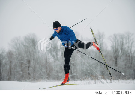 Male cross-country skier in blue jacket and black pants performs a dynamic maneuver on snow-covered trail with trees in the background during winter season Male cross-country skier in blue jacket and black pants performs a dynamic maneuver on snow-covered trail with trees in the background during winter season 140032938