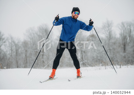 Male cross-country skier in blue jacket and black pants glides through snowy landscape, wearing sunglasses and ski poles, with trees in the background Male cross-country skier in blue jacket and black pants glides through snowy landscape, wearing sunglasses and ski poles, with trees in the background 140032944
