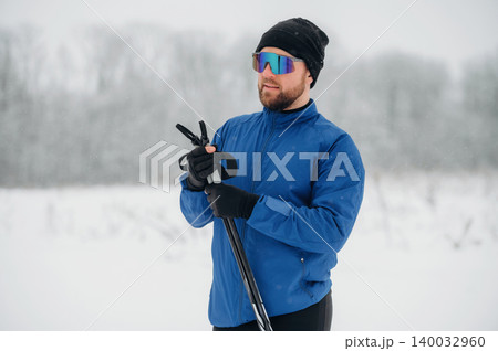 Male skier in blue jacket and sunglasses holds ski poles while standing in snowy landscape with blurred trees in the background during winter season 140032960