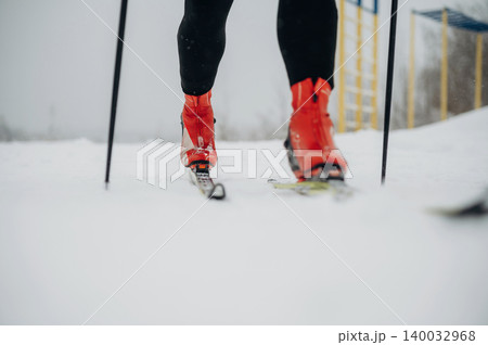 Skier wearing red boots and black pants glides on snow-covered terrain with ski poles, yellow structures visible in the background under overcast sky Skier wearing red boots and black pants glides on snow-covered terrain with ski poles, yellow structures visible in the background under overcast sky 140032968
