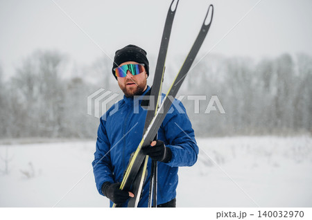 Male skier wearing blue jacket and sunglasses holds cross-country skis in snowy landscape with trees and falling snowflakes in the background Male skier wearing blue jacket and sunglasses holds cross-country skis in snowy landscape with trees and falling snowflakes in the background 140032970