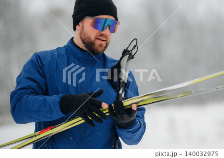 Male skier in blue jacket and black gloves adjusts ski poles while holding skis in snowy landscape with blurred trees in the background Male skier in blue jacket and black gloves adjusts ski poles while holding skis in snowy landscape with blurred trees in the background 140032975