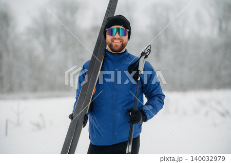 Male skier wearing blue jacket and sunglasses holds ski equipment in snowy landscape with trees and falling snowflakes in the background Male skier wearing blue jacket and sunglasses holds ski equipment in snowy landscape with trees and falling snowflakes in the background 140032979