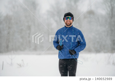 Male athlete wearing blue jacket and sunglasses stands in snowy landscape, snowflakes falling around him, winter trees blurred in the background Male athlete wearing blue jacket and sunglasses stands in snowy landscape, snowflakes falling around him, winter trees blurred in the background 140032989