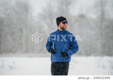 Male runner dressed in blue jacket and black pants jogs through snowy landscape wearing sunglasses and headphones during winter weather with trees in the background Male runner dressed in blue jacket and black pants jogs through snowy landscape wearing sunglasses and headphones during winter weather with trees in the background 140032990