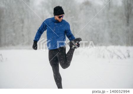 Male runner in blue long-sleeve shirt and black leggings stretches leg while jogging in snowy landscape with trees and falling snowflakes in the background Male runner in blue long-sleeve shirt and black leggings stretches leg while jogging in snowy landscape with trees and falling snowflakes in the background 140032994