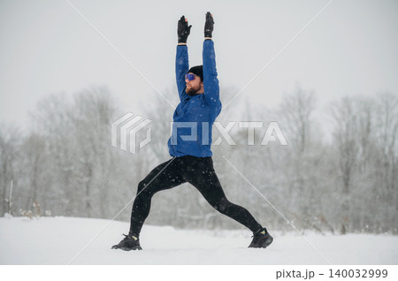 Male athlete performing yoga pose in snowy landscape, wearing blue jacket and black leggings, with snow-covered trees in the background 140032999