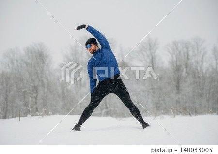 Male athlete stretching in snow-covered landscape during winter, wearing blue jacket and black leggings, with trees and snowfall in the background 140033005